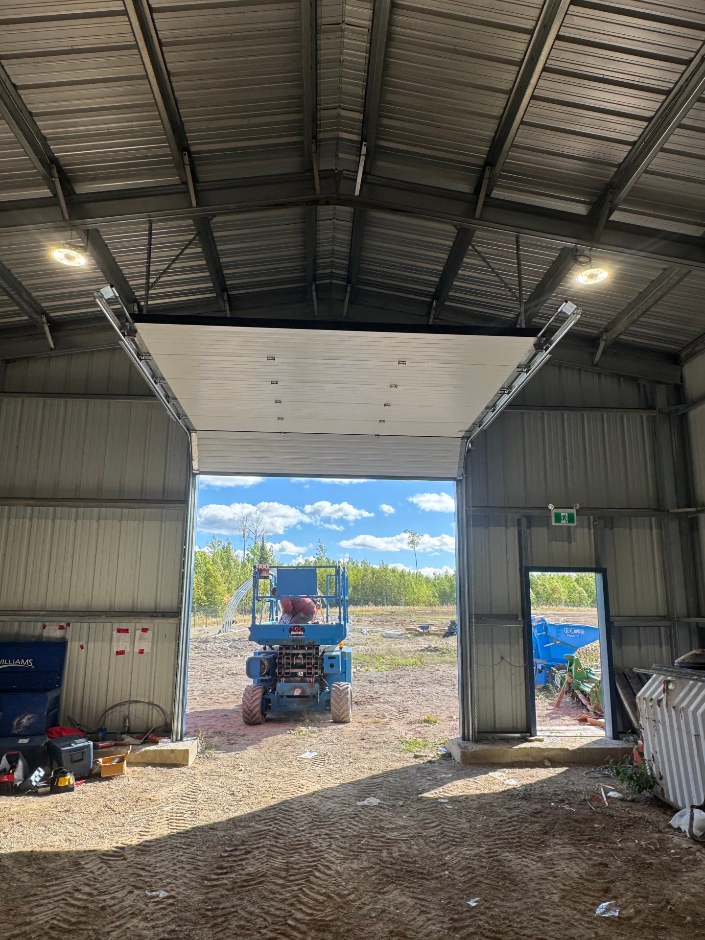 Inside a steel building with the overhead door open to trees outside and the ceiling lights on.