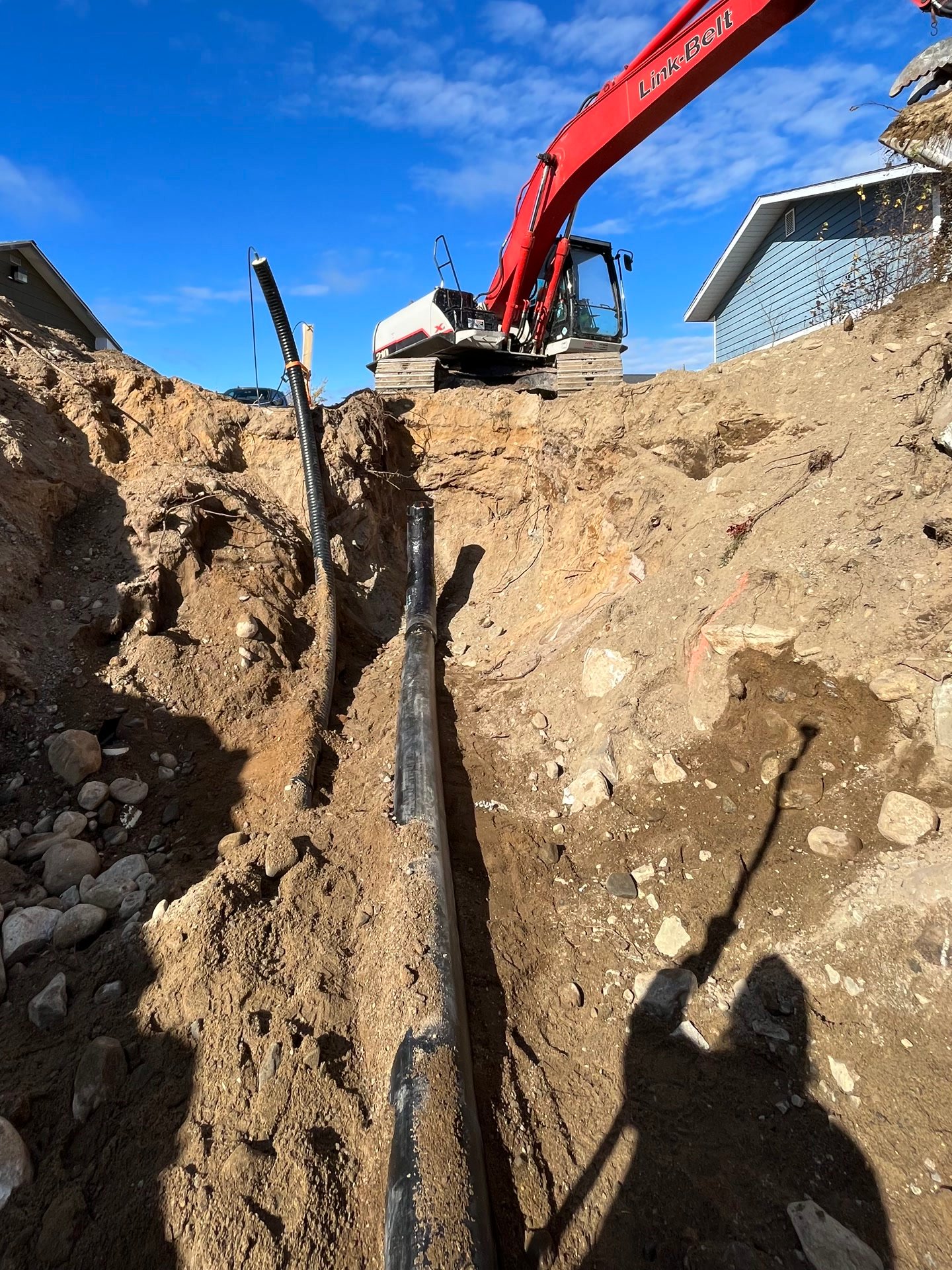 A service line being laid along the bottom of an open trench, with an excavator working above it under a clear sky.